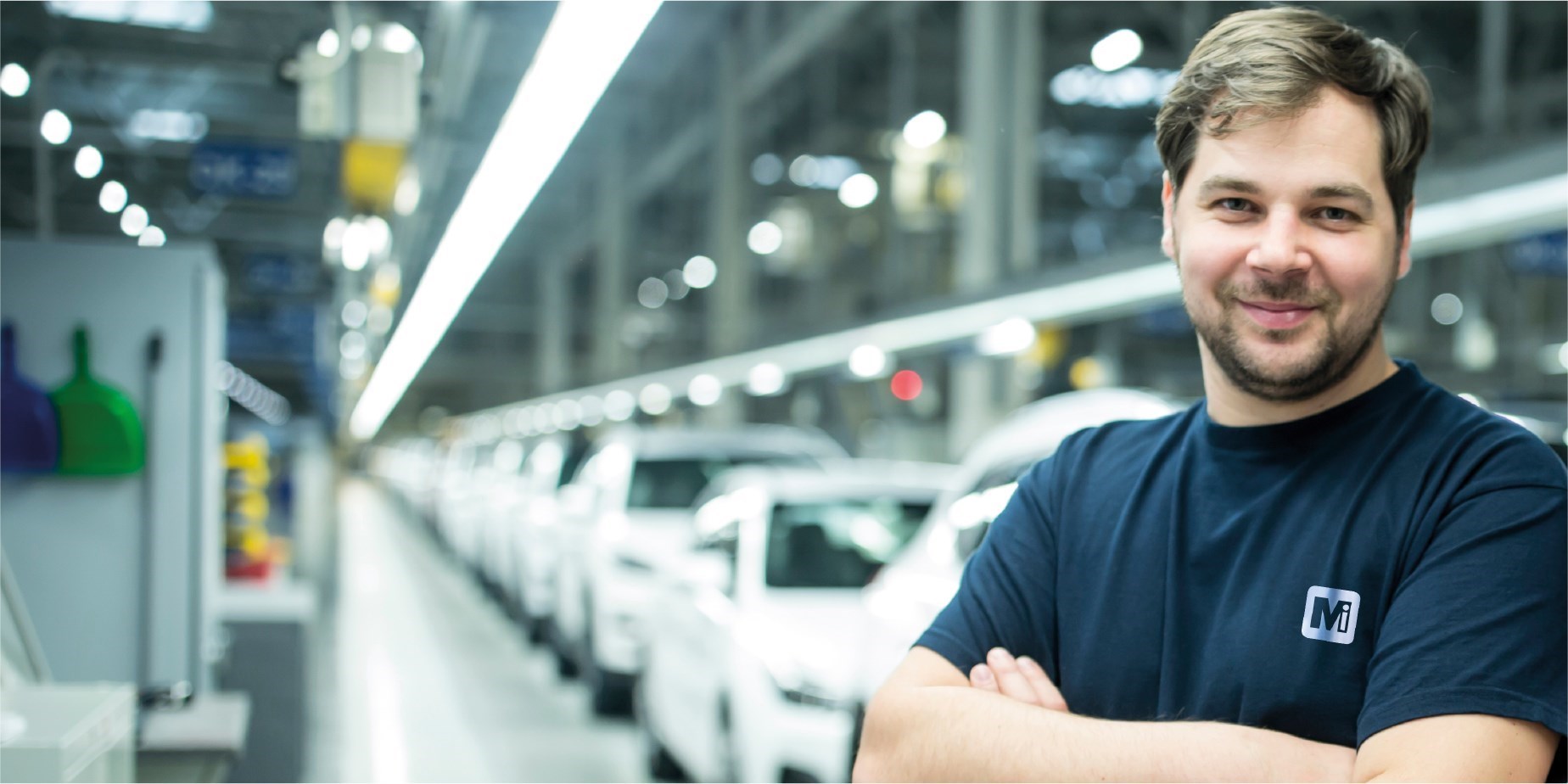 Man wearing a The Manufacturing Institute t-shirt with his arms folded standing in a car factory
