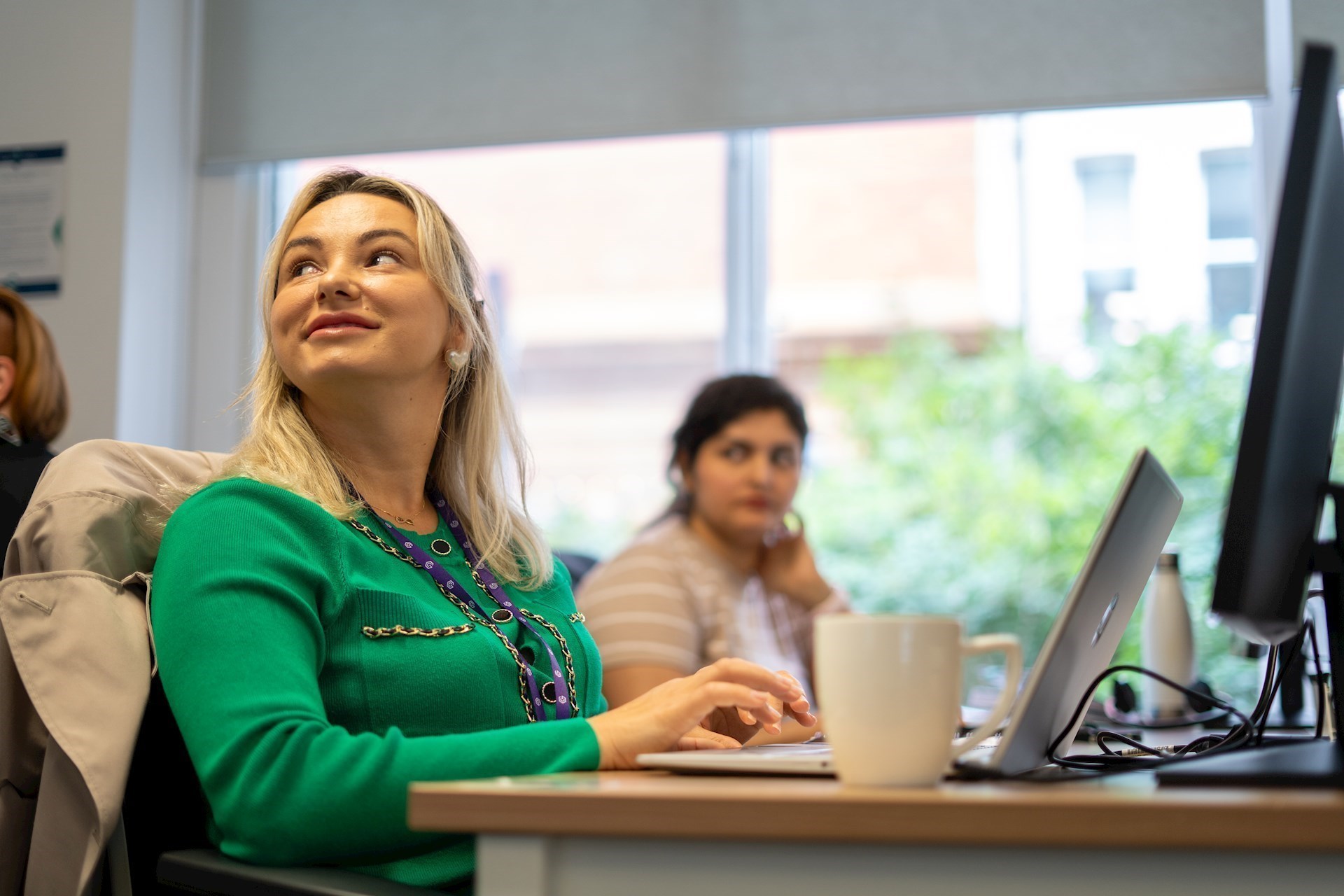 Two colleagues sitting at a desk in our office, the colleague closest is looking up to the left 