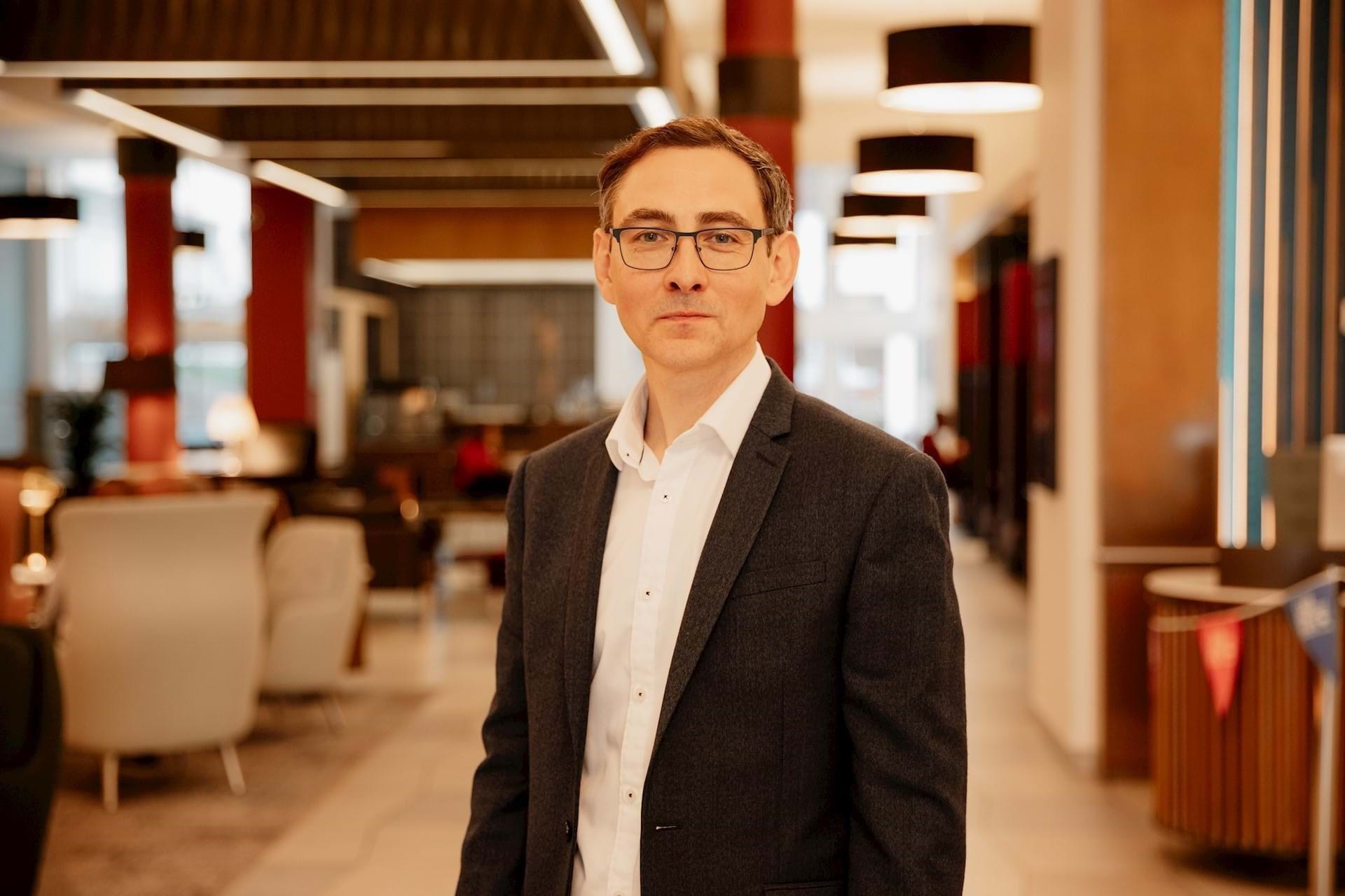 Richard Jeffery wearing a dark suit jacket with a white shirt standing in a modern office with a slightly blurred background