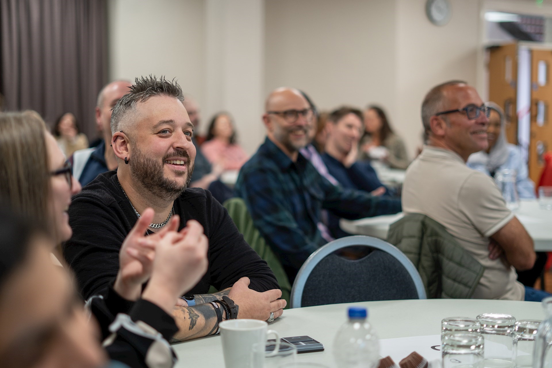 Colleague sitting at a table smiling with colleagues behind him slightly blurred