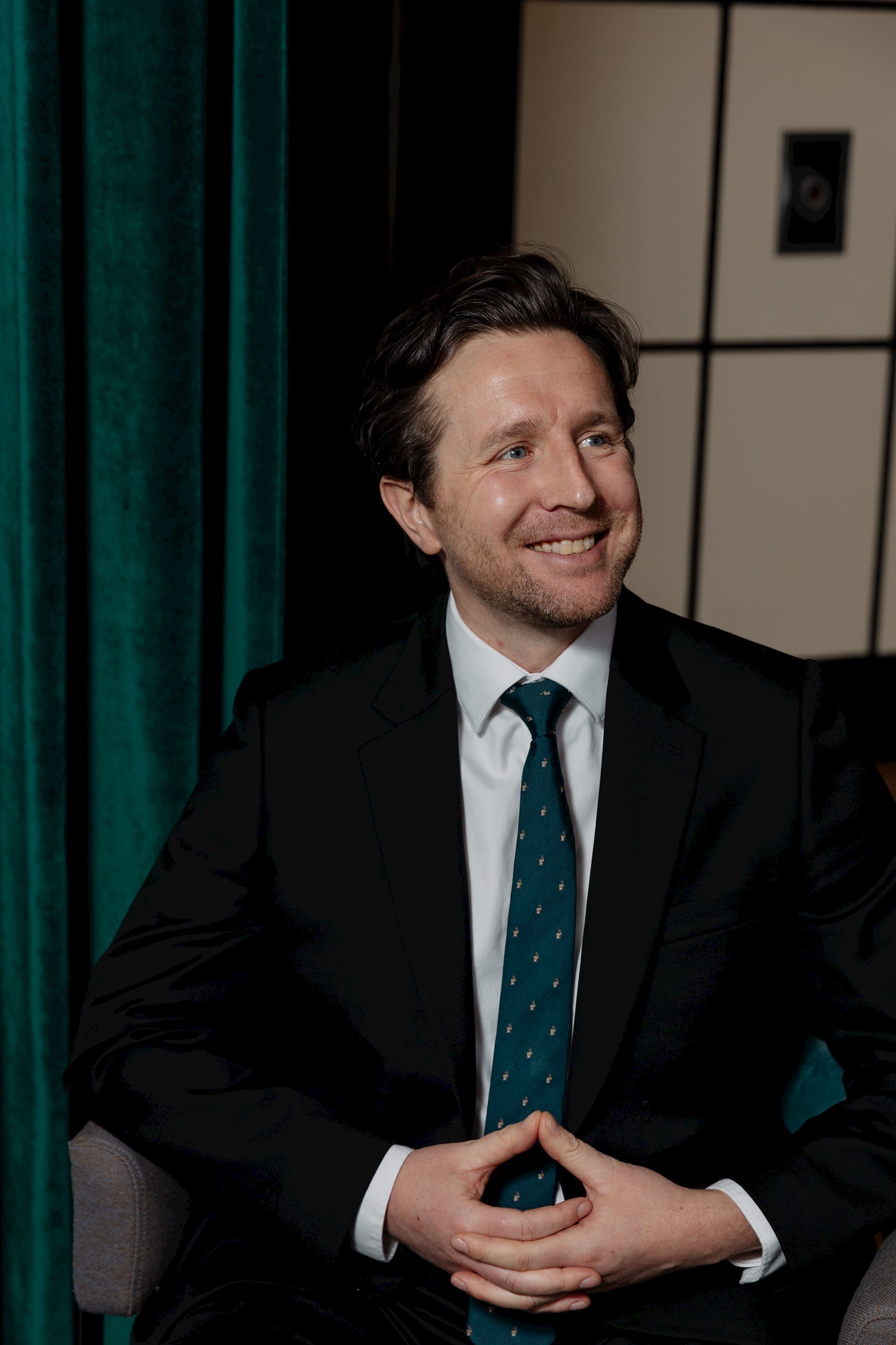 Joe Manning sitting and looking off to the side. He is wearing a black suit with a dark green tie and there is a green curtain in the background