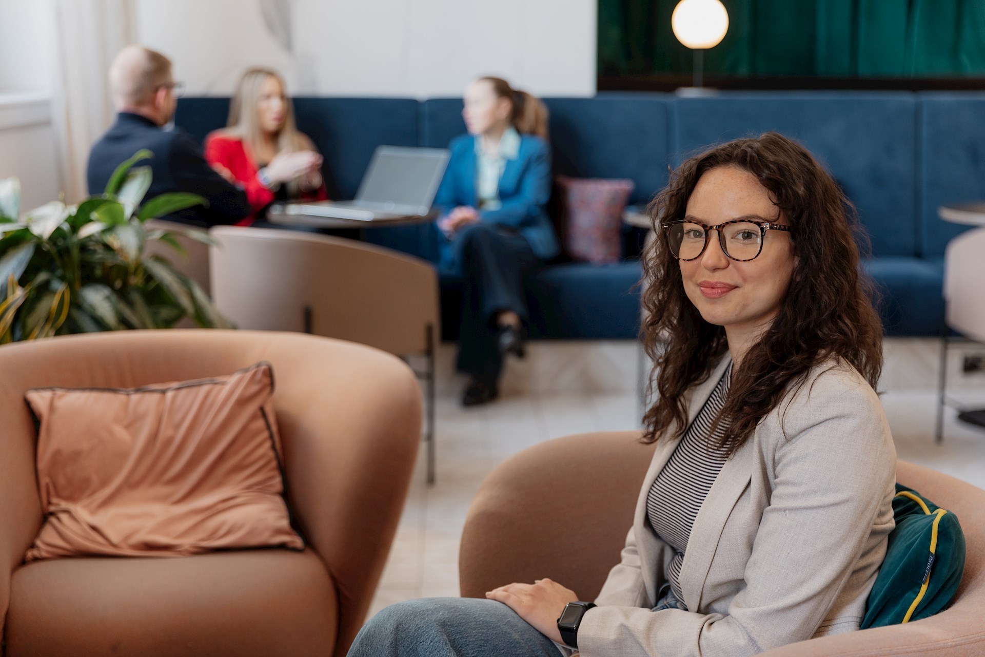 Young person sitting on a chair and looking to the left smiling towards the camera with colleagues blurred in the background