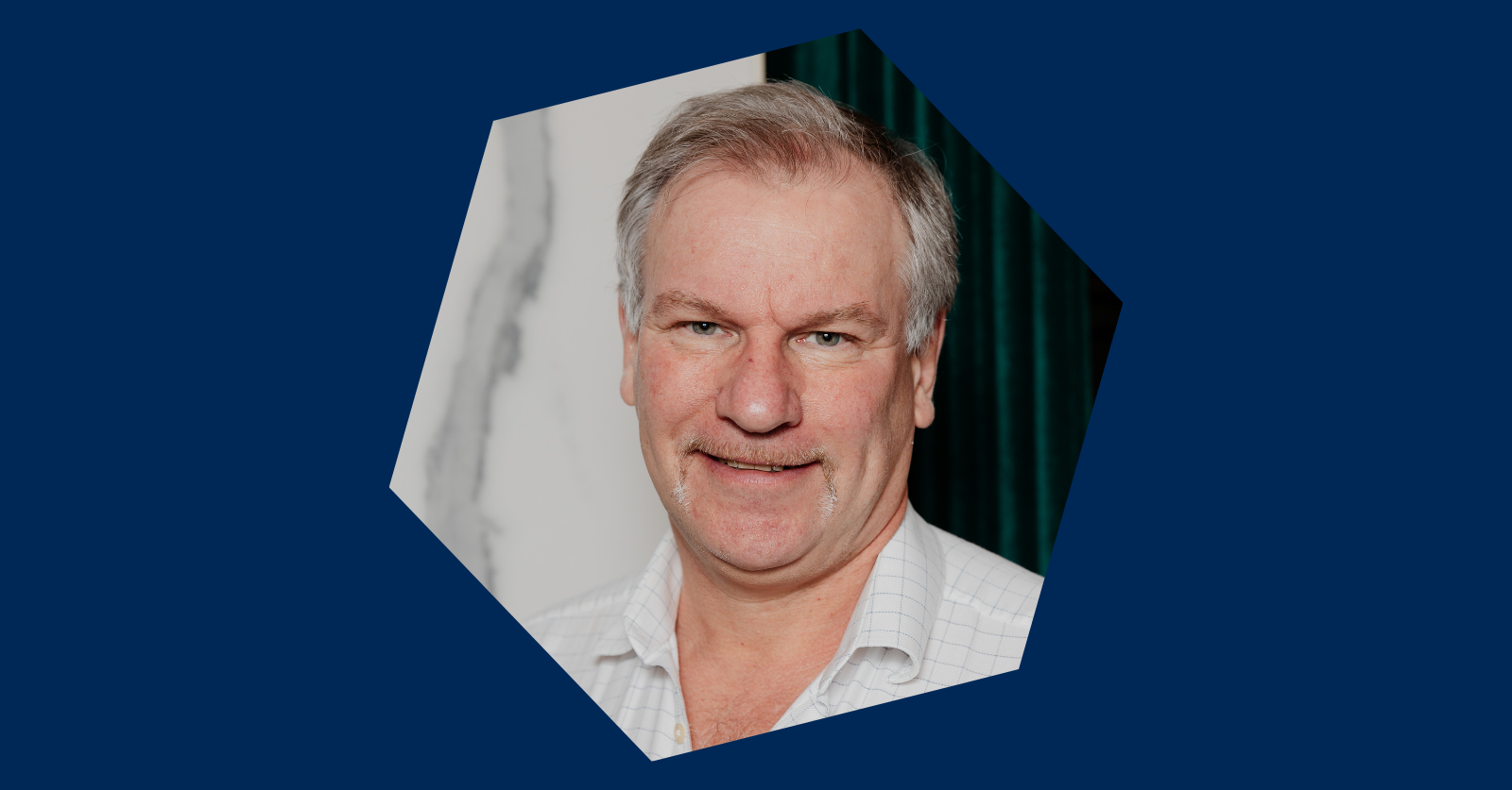 Professional headshot of Alain Dilworth wearing a white shirt in front of a marble wall. The image is in a hexagon shape on a dark blue background. 